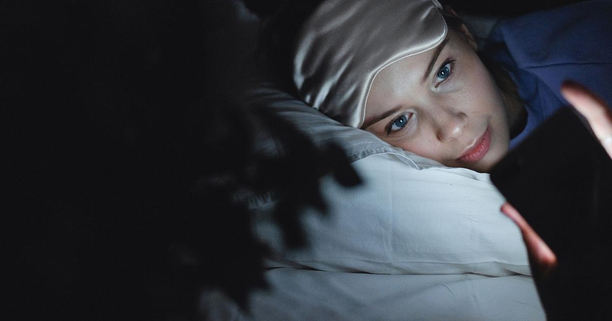 A woman lying in bed at night wearing a sleep mask, checking her phone by its glow instead of sleeping.